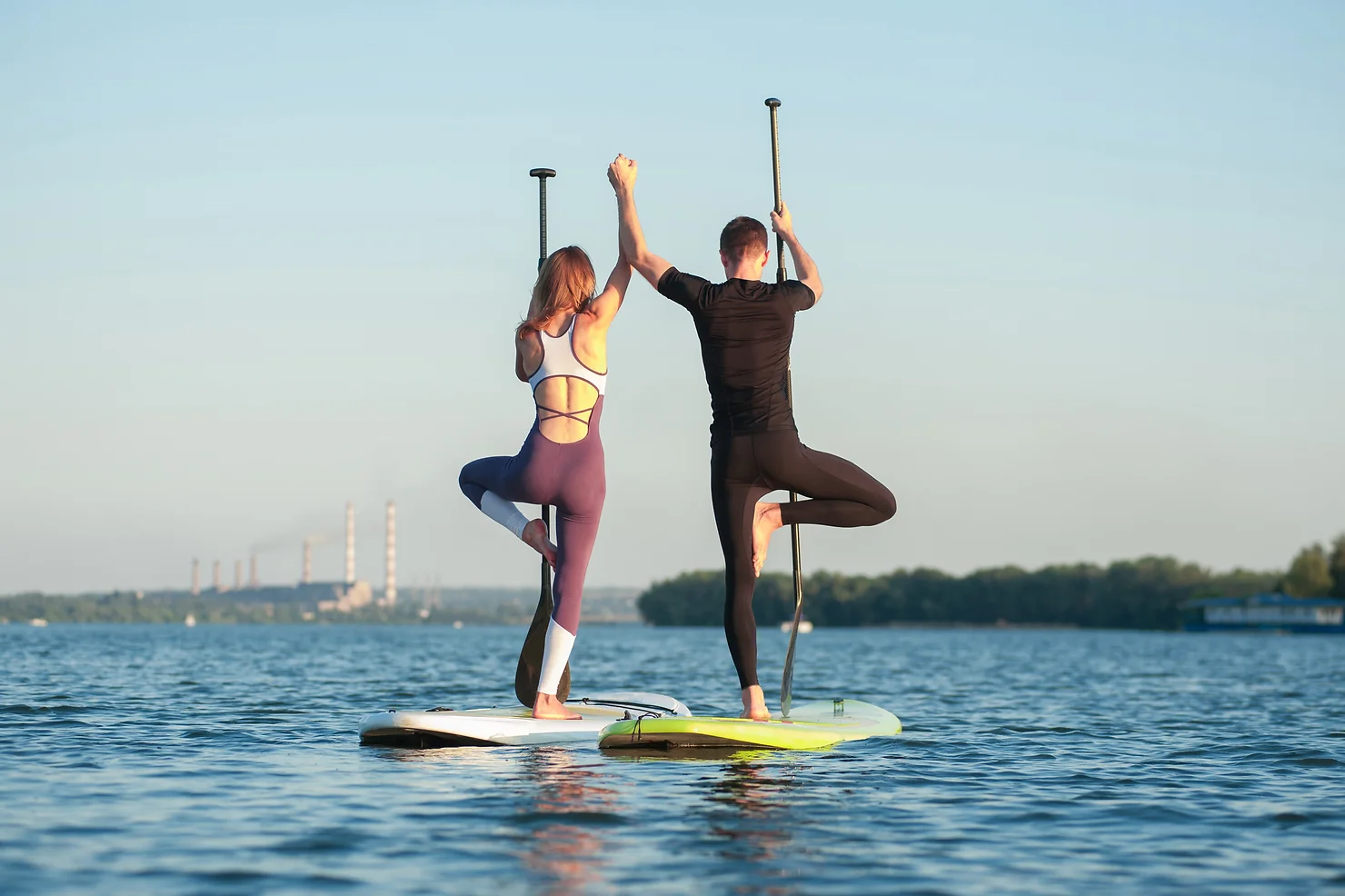 Auf diesem Bild stehe je ein Mann und eine Frau auf einem Standup Paddel. Sie balancieren auf einem Bein und sind auf einem See. Beide halten ein Paddel in der Hand und strecken diese Hand zur Seeseite. Dieses Bild steht für den Blogbeitrag: Wo stehen Sie?