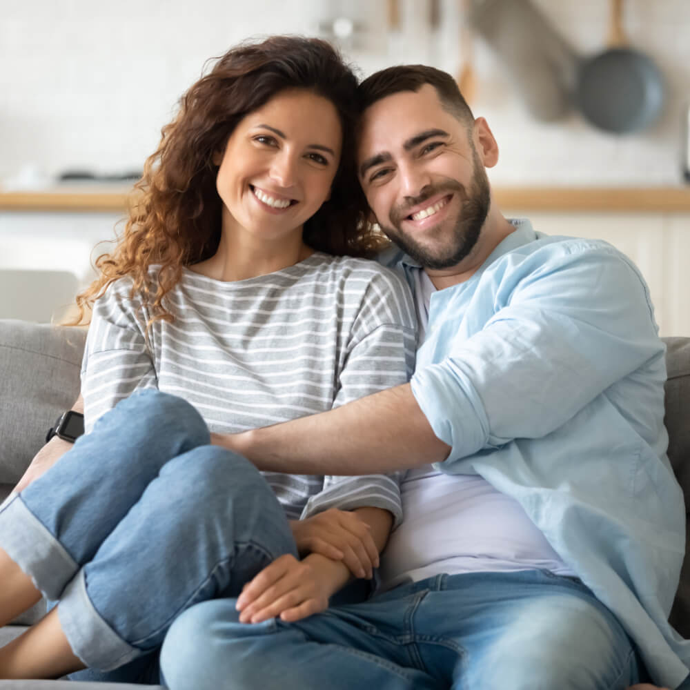 Ein Mann und eine Frau als Liebespaar sitzen auf einem Sofa und umarmen sich. Dieses Bild steht für das Angebot für Paartherapie. Dieses gilt auch für gleichgeschlechtliche Paare.