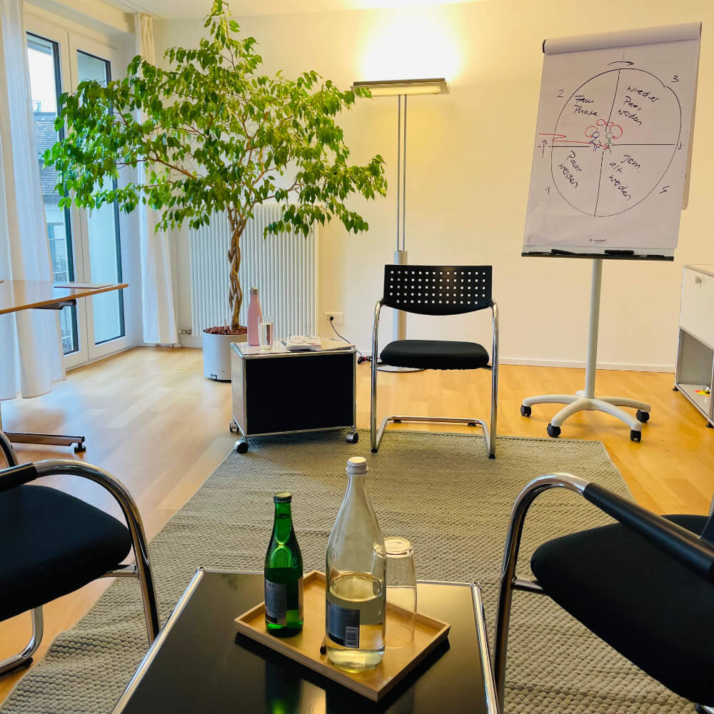 Praxis und Büro in Zürich von Nathalie Brady und Claude Keller. Einrichtung modern mit grossem Baum (grün) und einem lindgrünen Teppich und dunkelblauen Stühlen. Getränke stehen bereit. Der Raum ist hell und hat warmes Licht.